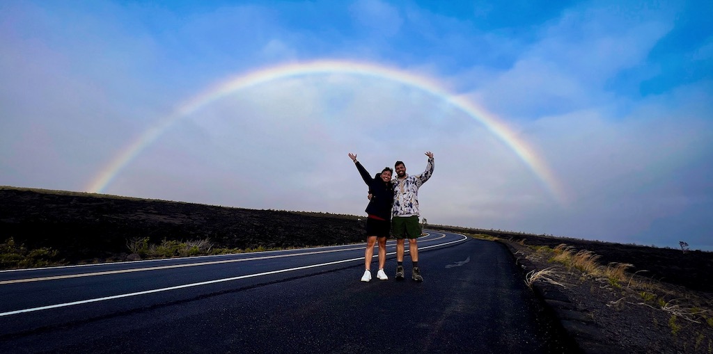AJ and his boyfriend with arms raised under a rainbow